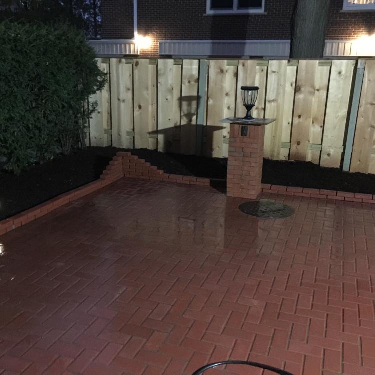 A backyard patio with a wooden fence, brick pillars, and outdoor lighting