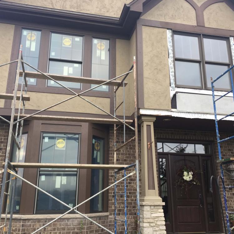 A two-story brick house with scaffolding surrounding the front facade. The house has a brown door, and the windows are being replaced